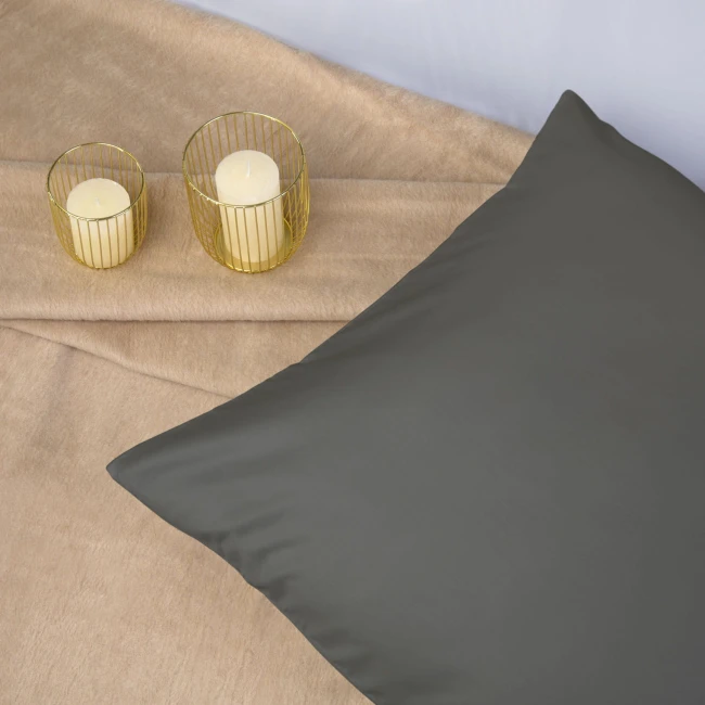 Poszewka satynowa w kolorze szarym 50x60 cm leży na beżowym tle. Na zdjęciu znajdują się również dwie świeczki w złotych osłonkach. Produkt jest w pełni widoczny, z gładką, błyszczącą powierzchnią materiału.
