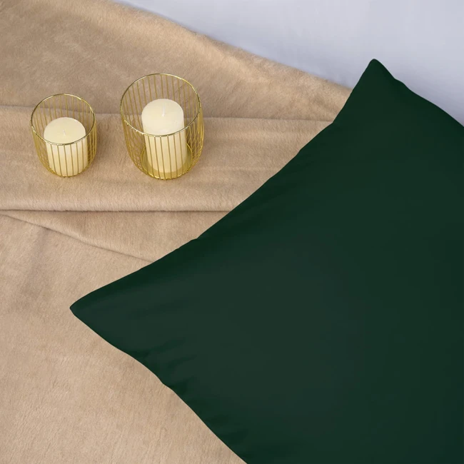 Poszewka satynowa 40x40 w kolorze butelkowej zieleni leży na beżowym, teksturowanym tle. W tle widoczne są dwa złote świeczniki z białymi świecami. Produkt ma gładką fakturę, bez widocznych napisów.