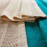 Ręcznik bawełniany 50x90 cm w kolorze ecru z ozdobnym bieżnikiem, leżący na tle turkusowego ręcznika. Widoczne detale tekstury, złożony w połowie, prezentujący jednocześnie strukturę materiału i wzór wplatania.