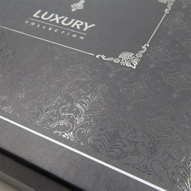 Opakowanie pościeli satynowej w kolorze ciemnoszarym z widocznym, wypukłym wzorem w stylu żakardowym. Na górze znajduje się napis LUXURY COLLECTION w eleganckim kroju, otoczony dekoracyjnym elementem. Opakowanie ma prostokątny kształt i jest zamknięte.