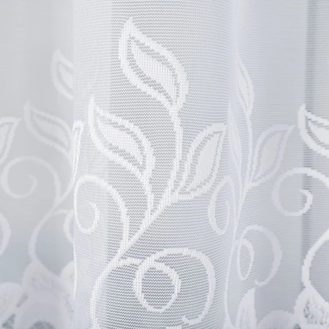 Biała firana żakardowa Bastia o wymiarach 500x160 cm, z delikatnym wzorem liści i spiral. Ujęcie zbliżeniowe, ukazujące fakturę i szczegóły wzoru. Podkreśla lekkość materiału, zawieszona, na jasnym tle.
