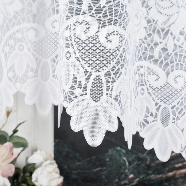 Firana gotowa Demetria żakardowa w kolorze białym o wymiarach 300x160 cm, z delikatnym wzorem florystycznym na dolnej krawędzi. Produkt wiszący, ukazany w zbliżeniu, z tłem w odcieniu ciemnej zieleni i różowych kwiatów.