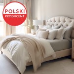 Prześcieradło jersey z elastanem w kolorze antracytowym, rozmiar 200x220 cm, leżące na łóżku. Obok poduszki w różnych odcieniach, tekstura gładka. W tle jasno oświetlona sypialnia z beżowymi firankami i lampą nocną.