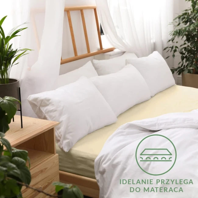 Prześcieradło jersey z gumką w kolorze kremowym, rozmiar 80x160, rozłożone na łóżku. Widoczne poduszki w białych poszewkach. Tło z ciemnego drewna oraz roślina w doniczce. Na materiale widoczna grafika z napisem Idealnie przylega do materaca.