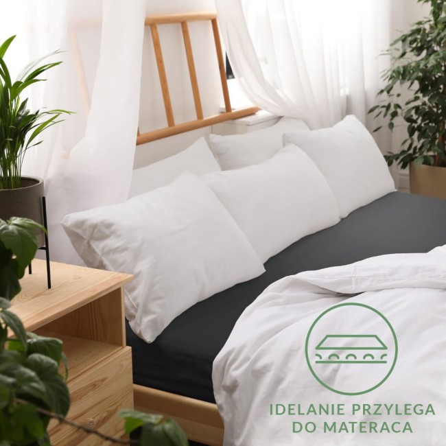 Czarne prześcieradło jersey z gumką 200x220, rozłożone na łóżku, z białymi poduszkami w tle. Widoczna faktura materiału, idealnie dopasowane do materaca. Otoczenie jasne, z roślinnością w donicy oraz drewnianymi meblami.
