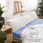 Prześcieradło jersey z gumką w kolorze niebieskim o wymiarach 160x200 cm, rozłożone na łóżku. W tle widoczne białe poduszki i lity stelaż łóżka.