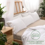 Prześcieradło jersey z gumką 90x200 w kolorze beżowym, rozłożone na łóżku, z widocznym białym zestawem poduszek w tle. Tło z jasnymi zasłonami i rośliną doniczkową. Widoczny napis Idealnie przylega do materaca.