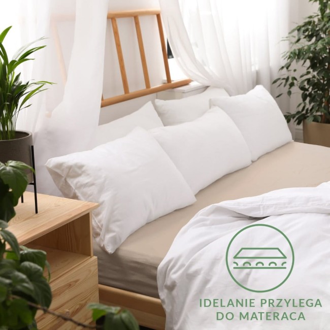 Prześcieradło jersey z gumką 180x200 w kolorze beżowym, leżące na łóżku z białymi poduszkami. Tło z jasnymi zasłonami i drewnianym stelażem łóżka oraz rośliną w doniczce na stojaku. Widoczne logo z hasłem Idealnie przylega do materaca.