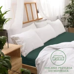 Prześcieradło jersey z gumką 120x200 w kolorze butelkowej zieleni, rozłożone na łóżku z białą pościelą. Widoczna guma brzegowa. W tle drewniane elementy, roślinność i okno z zasłonami. Elegancka aranżacja sypialni, zbliżenie na materiał.