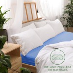 Prześcieradło jersey z gumką 180x200 w kolorze chaber, rozłożone na łóżku z białymi poduszkami. Kąt ujęcia frontalny, ukazujący sypialnię z naturalnym światłem. Widoczna informacja Idealnie przylega do materaca. Tło jasne, z drewnianymi elementami.