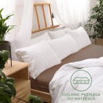 Prześcieradło jersey z gumką 90x200 w kolorze czekoladowym na łóżku, z białymi poduszkami ułożonymi wzdłuż. Produkt leży na rozłożonym materacu, z delikatnym białym prześcieradłem częściowo odsłoniętym. W tle widać rośliny oraz drewniane elementy mebli.