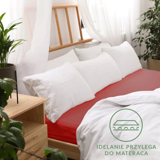 Prześcieradło jersey z gumką w kolorze czerwonym, rozmiar 90x200, leżące na łóżku z białymi poduszkami. Widoczne jest logo oraz tekst IDEALNIE PRZYLEGA DO MATERACA. Tło stanowi jasne wnętrze z roślinnością i drewnianymi meblami.