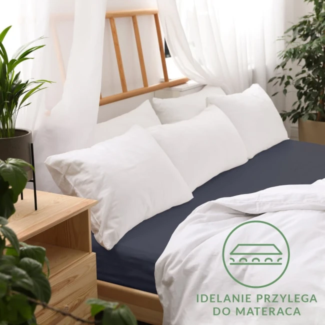 Prześcieradło jersey z gumką w kolorze ciemnego granatu leży na łóżku z białą pościelą i poduszkami. Ujęcie z góry pokazuje gładką teksturę materiału oraz gumkę, która zapewnia idealne dopasowanie do materaca.