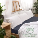Prześcieradło jersey z gumką 140x200 w kolorze ciemnym granatowym, leżące na łóżku z białą pościelą i poduszkami. W tle widoczne drewniane łóżko i rośliny. Prześcieradło idealnie przylega do materaca, materiał gładki, bez wzorów.