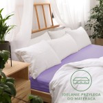 Prześcieradło jersey z gumką 180x200 w kolorze jasnym fiolecie leży na łóżku z białą pościelą i poduszkami. Kąt ujęcia z boku pokazuje szczegóły konstrukcji oraz fakturę materiału.