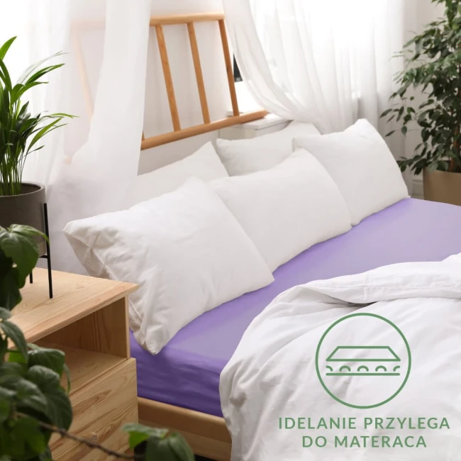 Prześcieradło jersey z gumką 120x200 w kolorze jasnym fiolecie, rozłożone na łóżku. Widoczne białe poduszki i miękka narzuta. Tło naturalne z drewnianymi elementami oraz roślinnością, z logo „Idealnie przylega do materaca” w rogu.
