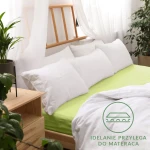 Prześcieradło jersey z gumką w kolorze limonkowym 140x200, rozłożone na łóżku z białymi poduszkami. Widoczne są gładkie, lekko teksturowane powierzchnie. Tło jasne, z rośliną i drewnianymi meblami w aranżacji sypialni.