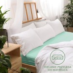 Prześcieradło jersey z gumką 140x200 w kolorze morskim, rozłożone na łóżku z białymi poduszkami. Ujęcie z boku, pokazujące szczelne przyleganie do materaca. Tło składa się z naturalnych elementów, takich jak rośliny i drewniane meble.
