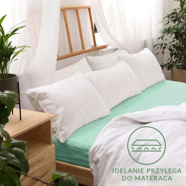 Prześcieradło jersey z gumką 140x200 w kolorze morskim, rozłożone na łóżku z białymi poduszkami. Ujęcie z boku, pokazujące szczelne przyleganie do materaca. Tło składa się z naturalnych elementów, takich jak rośliny i drewniane meble.