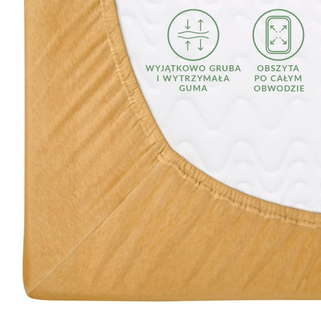 Prześcieradło jersey z gumką 120x200 w kolorze musztardowym, leżące na białym tle. Zbliżenie na róg, pokazujące wyjątkowo grubą i wytrzymałą gumę oraz obszytą tkaniną. Widoczne napisy dotyczące cech produktu, na delikatnej teksturze materiału.