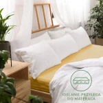 Prześcieradło jersey z gumką 160x200 w kolorze musztardowym, rozłożone na łóżku. Widoczne białe poduszki w tle oraz drewniana rama łóżka. Tło jasne, z zasłonami i rośliną. Na prześcieradle widoczny nadruk „IDEALNIE PRZYLEGA DO MATERACA”.