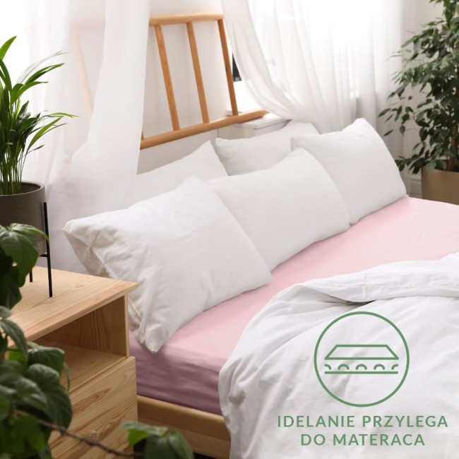 Prześcieradło jersey z gumką w kolorze różu kwarcowego, rozmiar 120x200, rozłożone na łóżku z białymi poduszkami. Ujęcie pokazuje je na tle jasnych zasłon i drewnianego wezgłowia. Widoczne logo i napis idealnie przylega do materaca.