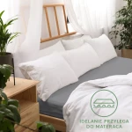 Prześcieradło jersey z gumką 120x200 w kolorze szarym leży na łóżku, obok białych poduszek. Widoczny jest detal gumki, która zapewnia idealne przyleganie do materaca. Tło jest jasne z naturalnymi akcentami roślinnymi.