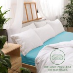 Prześcieradło jersey z gumką 90x200 w kolorze turkusowym, rozłożone na łóżku z białą pościelą i poduszkami. W tle widoczny drewniany zagłówek oraz rośliny doniczkowe. Na prześcieradle logo z napisem idealnie przylega do materaca.