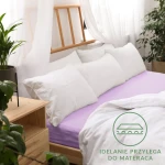 Prześcieradło jersey z gumką 160x200 w kolorze wrzosowym, rozłożone na łóżku z białą pościelą i poduszkami. Ujęcie z góry, ukazuje fakturę materiału oraz wnętrze z roślinami i drewnianym stolikiem.