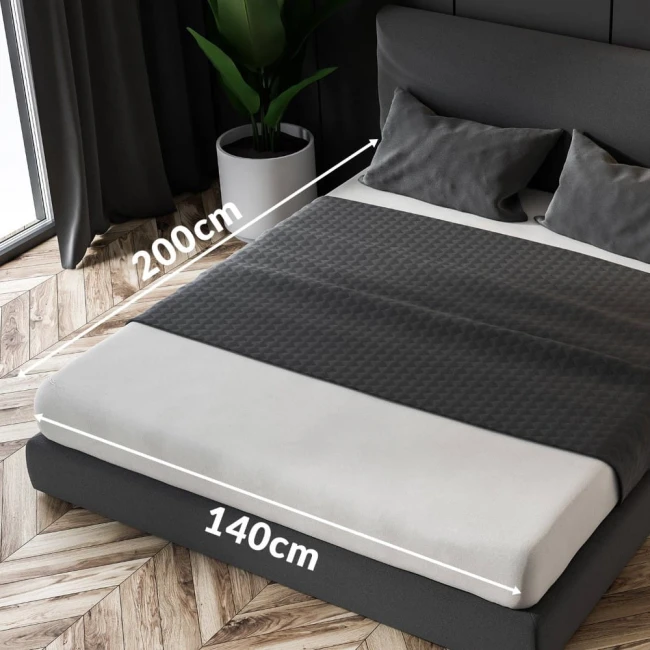 Prześcieradło frotte z gumką w kolorze popielatym, o wymiarach 140x200 cm. Ułożone na łóżku, ozdobione białą lamówką. W tle widoczne poduszki o szarym materiale oraz roślina w doniczce. Zdjęcie w kącie z góry, ukazujące całe łóżko.