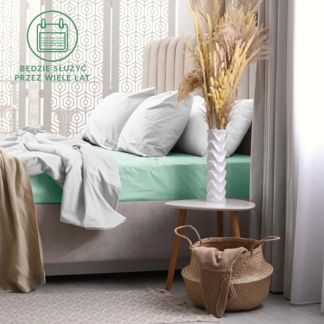 Prześcieradło frotte z gumką 180x200 w kolorze morskim leży na łóżku, nakryte białą pościelą. Produkt jest rozłożony na materacu, doskonale dopasowany do krawędzi. W tle widoczny jest stolik nocny z białą wazonem i suszonymi kwiatami.
