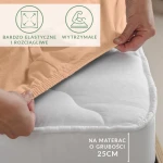 Prześcieradło jersey z elastanem w kolorze różowym, z gumką, leży na białym materacu o grubości 25 cm. W widoku, ręka trzyma jedną krawędź prześcieradła, które jest rozciągliwe i dopasowuje się do kształtu materaca.