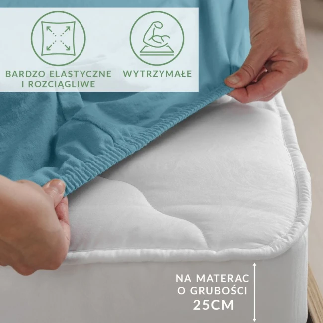Prześcieradło jersey z elastanem w kolorze błękitnym, rozłożone na materacu, z widoczną gumką. Kąt ujęcia z boku, ukazujący elastyczność materiału. Na materacu o grubości 25 cm. W tle naturalne wykończenie podłogi.