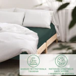 Prześcieradło satynowe bez gumki w kolorze butelkowej zieleni, rozłożone na łóżku z białą pościelą. Kąt ujęcia frontalny, z widocznym drewnianym stelażem. Gładka faktura materiału podkreśla jego wytrzymałość i miękkość. W tle jasne zasłony i poduszki.