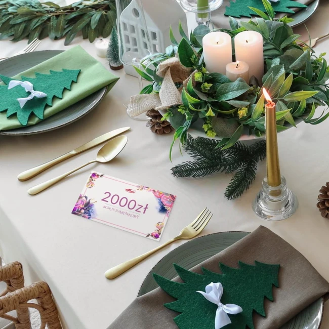 Karta podarunkowa o wartości 2000 zł leżąca na eleganckim stole. Stół z obrusami w odcieniach beżu i zieleni, otoczony dekoracjami, w tym świecami i gałązkami.