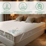 Ochraniacz na materac 60x120 cm w kolorze białym, leżący na materacu, z widocznymi poduszkami na łóżku. Na powierzchni ochraniacza widoczne rozlane woda z szklanki, co podkreśla jego nieprzemakalne właściwości. Proste tło z ciepłym oświetleniem wnętrza.