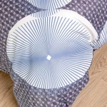 Biała pościel satynowa w rozmiarze 200x220 cm z niebieskimi okręgami i liniami. Kąt ujęcia: z góry, ukazujący wzór i fakturę. Tło jasne, podłoga drewniana. Pościel rozłożona na powierzchni.