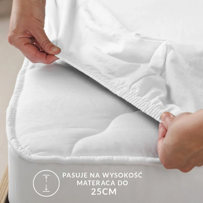 Ochronne prześcieradło z gumką w kolorze beżowym, pasujące na materac do 25 cm wysokości. Zbliżenie na rękę trzymającą brzeg prześcieradła, które przylega do materaca. Faktura materiału delikatna, gładka, bez wzorów. Tło neutralne.