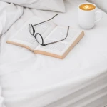 Prześcieradło bawełniane hotelowe białe 160x260 GŁADKIE leży na łóżku. Na nim znajduje się otwarta książka oraz okulary w kolorze czarnym.