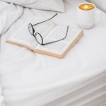 Prześcieradło bawełniane hotelowe białe 160x240 GŁADKIE rozłożone na łóżku, z książką otwartą i okularami położonymi na stronach. W tle filiżanka z kawą. Całość na jasnym tle pościeli, w neutralnych kolorach.