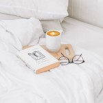Prześcieradło bawełniane hotelowe białe 160x200, gładkie, leżące na łóżku. Obok książka i filiżanka z kawą na drewnianej desce. Tło białe, jasno oświetlone, tworzące przytulną atmosferę. Bliższe ujęcie detali tkaniny oraz aranżacji.