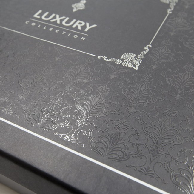Czarna, elegancka opakowanie z tłoczonym wzorem roślinnym w odcieniach srebra, z napisem LUXURY COLLECTION. Widoczne detale tekstury i wzoru, kąt ujęcia z góry, zbliżenie na dekoracyjne elementy opakowania.