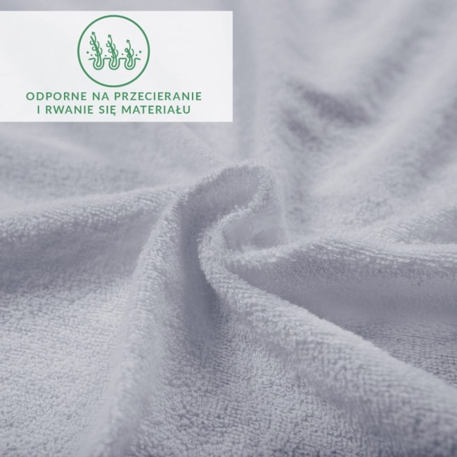 Prześcieradło frotte z gumką w kolorze popielatym, o wymiarach 180x200 cm, z bliska ukazuje miękką, teksturowaną powierzchnię. Widoczne zawinięcia materiału podkreślają jego elastyczność. Tło jest jednolite, co skupia uwagę na detalu tkaniny.
