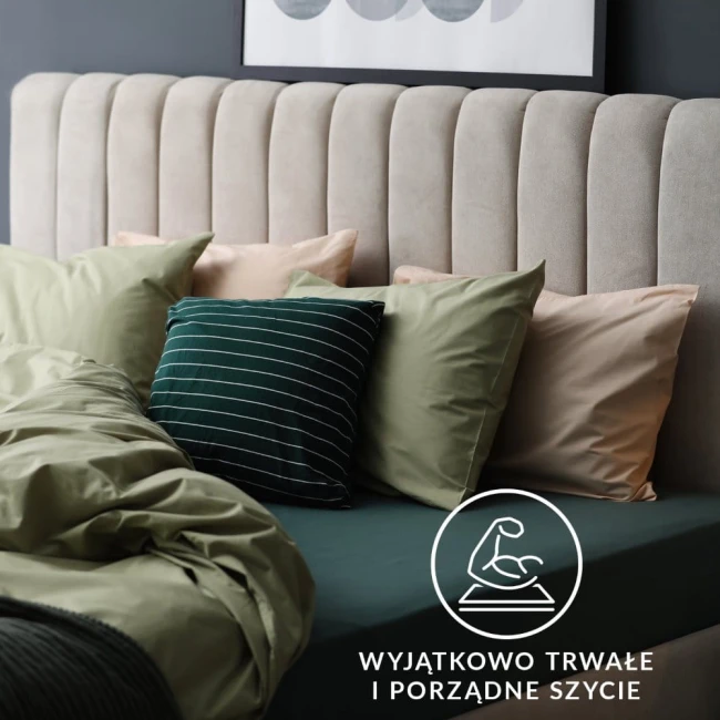 Prześcieradło jersey z gumką 90x200 w kolorze białym leży na łóżku, przykryte miękką narzutą. W tle widać headboard z pionowymi przeszyciami oraz różnokolorowe poduszki. Widoczna jest również etykieta z napisem o trwałości i jakości szycia.