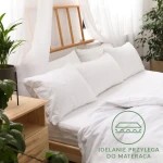 Prześcieradło jersey z gumką 90x200 w kolorze białym, rozłożone na łóżku z jasnym, drewnianym stelażem. Zastosowana gumka zapewnia idealne przyleganie. W tle białe poduszki oraz rośliny, całość na jasnym tle.