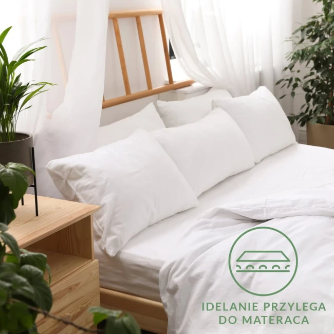 Prześcieradło jersey z gumką 90x200 w kolorze białym, rozłożone na łóżku z jasnym, drewnianym stelażem. Zastosowana gumka zapewnia idealne przyleganie. W tle białe poduszki oraz rośliny, całość na jasnym tle.
