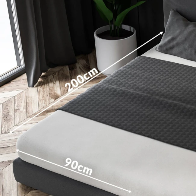 Prześcieradło jersey z gumką w kolorze białym, rozmiar 90x200 cm, leży na łóżku. Widoczna faktura materiału oraz oznaczenia wymiarów, w tle roślina w doniczce i zasłony. Prześcieradło idealnie dopasowane do materaca.
