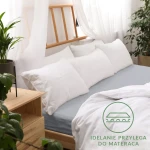 Prześcieradło jersey z gumką w kolorze popielatym o wymiarach 120x200 cm, rozłożone na łóżku, idealnie dopasowane do materaca. W tle biała pościel i poduszki na łóżku oraz drewniana rama. Widoczny napis IDEALNIE PRZYLEGA DO MATERACA w zielonym kole.