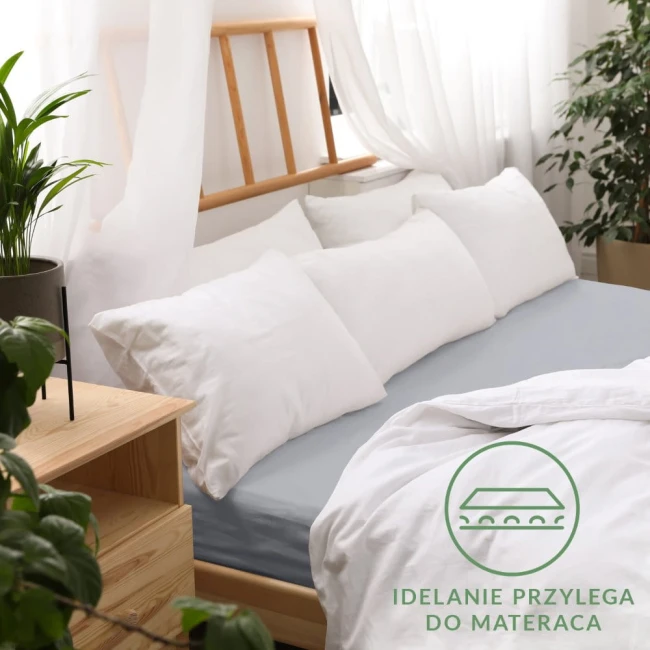 Prześcieradło jersey z gumką w kolorze popielatym o wymiarach 120x200 cm, rozłożone na łóżku, idealnie dopasowane do materaca. W tle biała pościel i poduszki na łóżku oraz drewniana rama. Widoczny napis IDEALNIE PRZYLEGA DO MATERACA w zielonym kole.