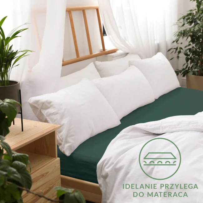 Prześcieradło jersey z gumką w kolorze zieleni butelkowej, rozmiar 200x220 cm, idealnie dopasowane do materaca. Położone na łóżku z białymi poduszkami i narzutą, w otoczeniu rośliny doniczkowej i drewnianego stolika.